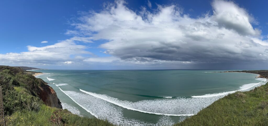 Photo of Anglesea from the main beach lookout. The image looks over Point Roadknight beach/ Soapy Rocks and Anglesea Main Beach of the Ocean, beach and bush on a calm day with waves and storm coming in.