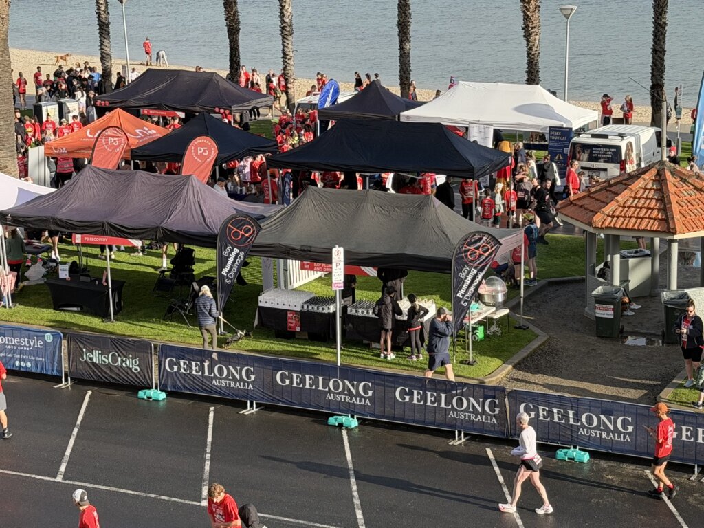 Run4Geelong event, showing people and tents on the Geelong water front with easter beach water in the background and Palm trees. Sponsor tents with Bay Plumbing & Drainage and other sponsors.